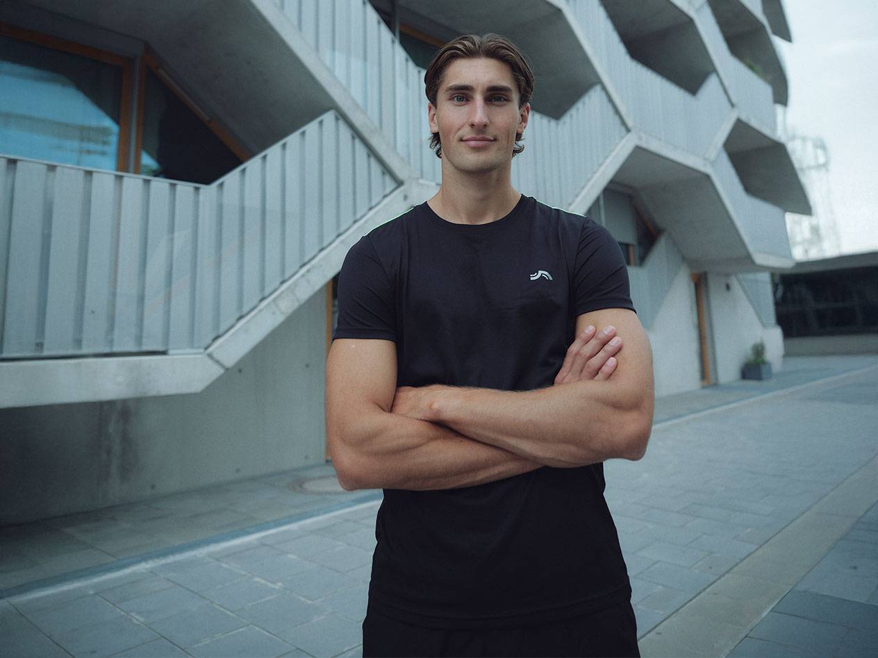 Homme en t-shirt noir avec logo et short noir, debout à l'extérieur les bras croisés.