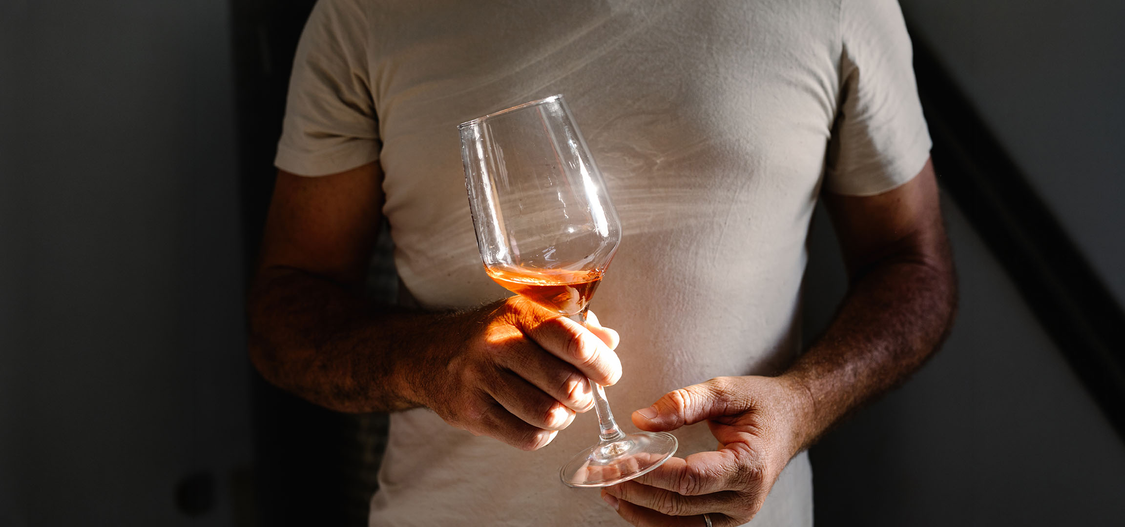 Homme tenant un verre de vin rosé, baigné de soleil, sur fond sombre.