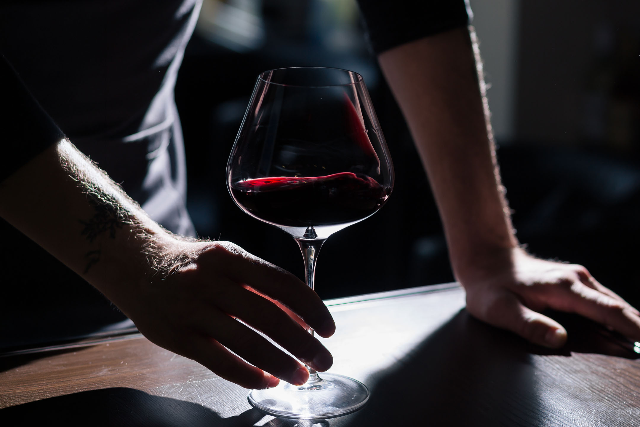 Mains tatouées d'un homme tenant un verre de vin rouge sur une table en bois.