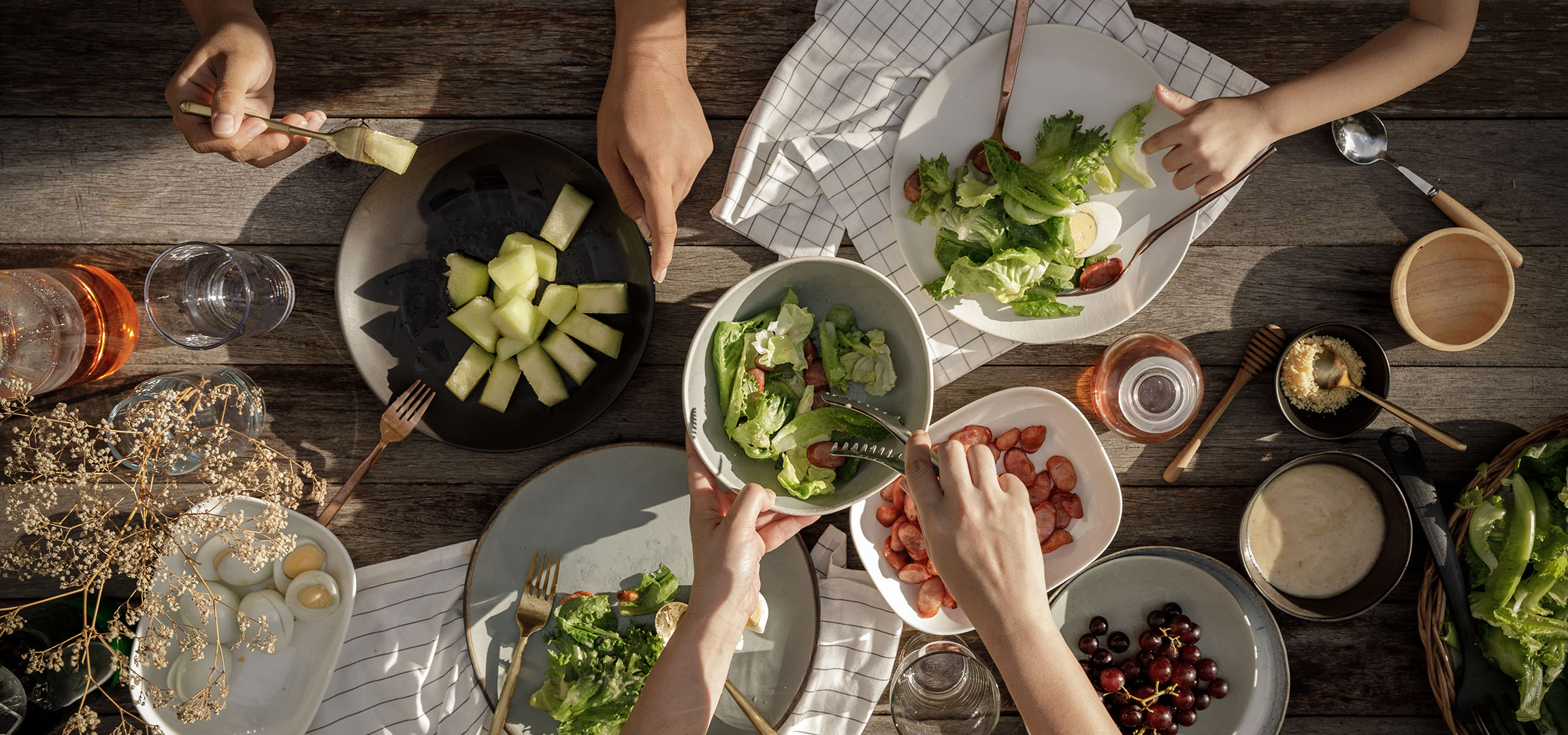 Vue de dessus d'une table avec salade, saucisses et melon, des personnes servant à manger.