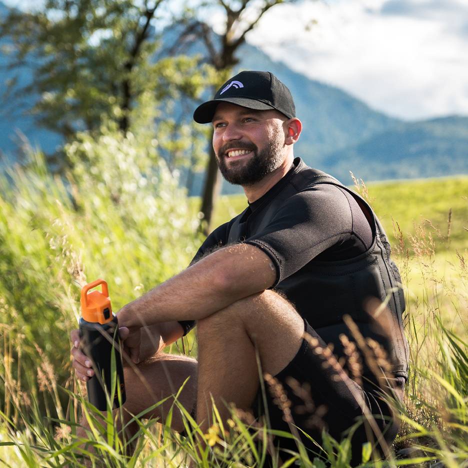 Homme souriant avec casquette noire et gilet en néoprène, tenant une bouteille d'eau orange.