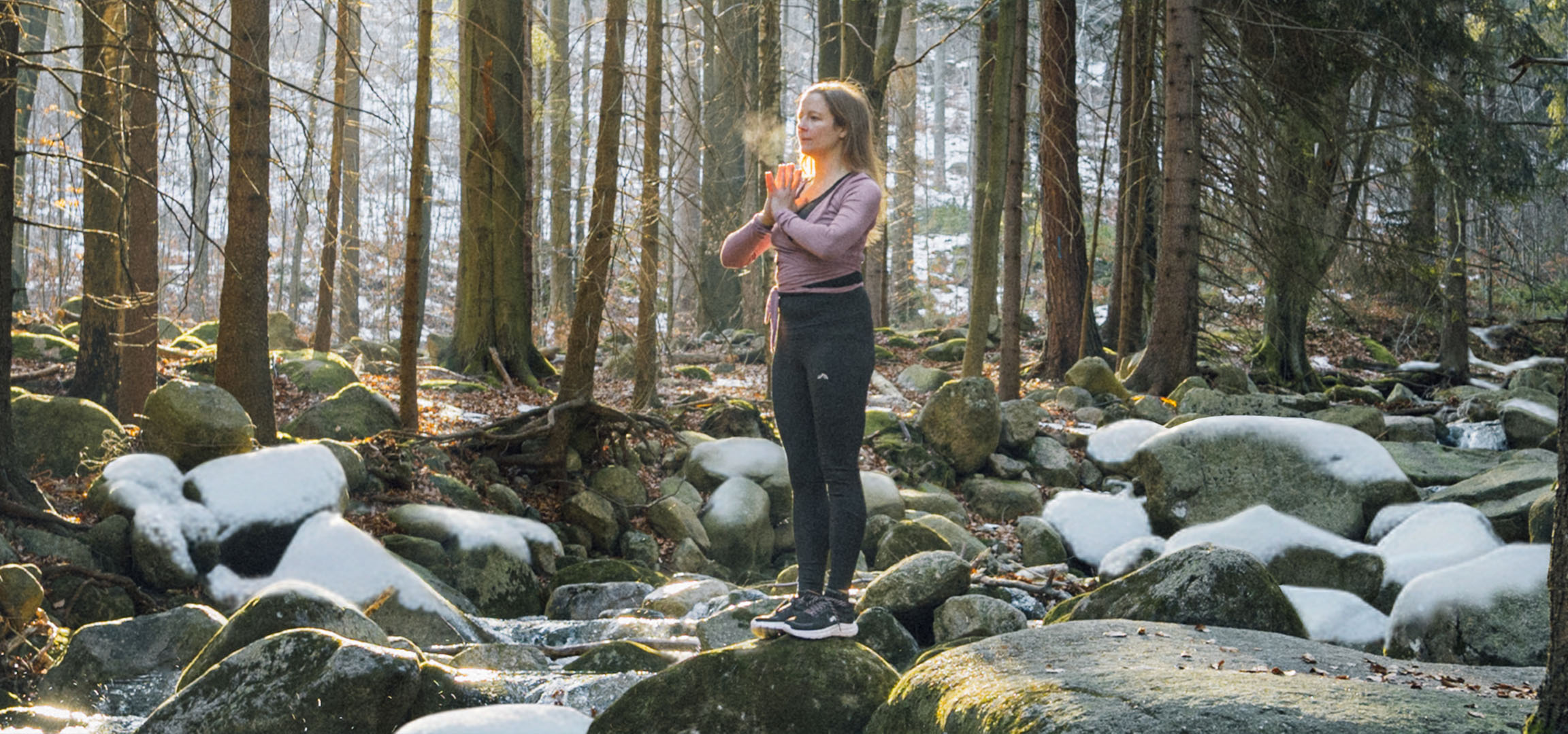Femme en tenue de sport faisant du yoga dans une forêt enneigée avec un ruisseau.