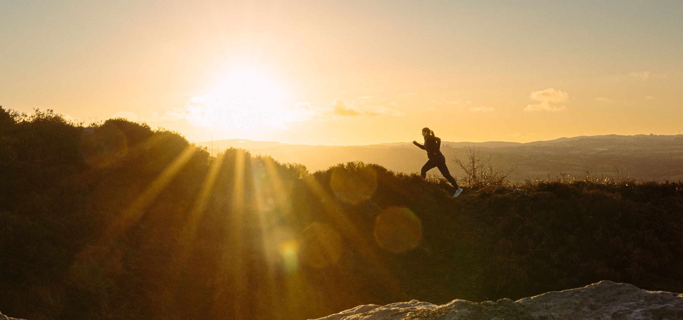Femme courant sur une colline au coucher du soleil, avec des rayons de soleil traversant le paysage.