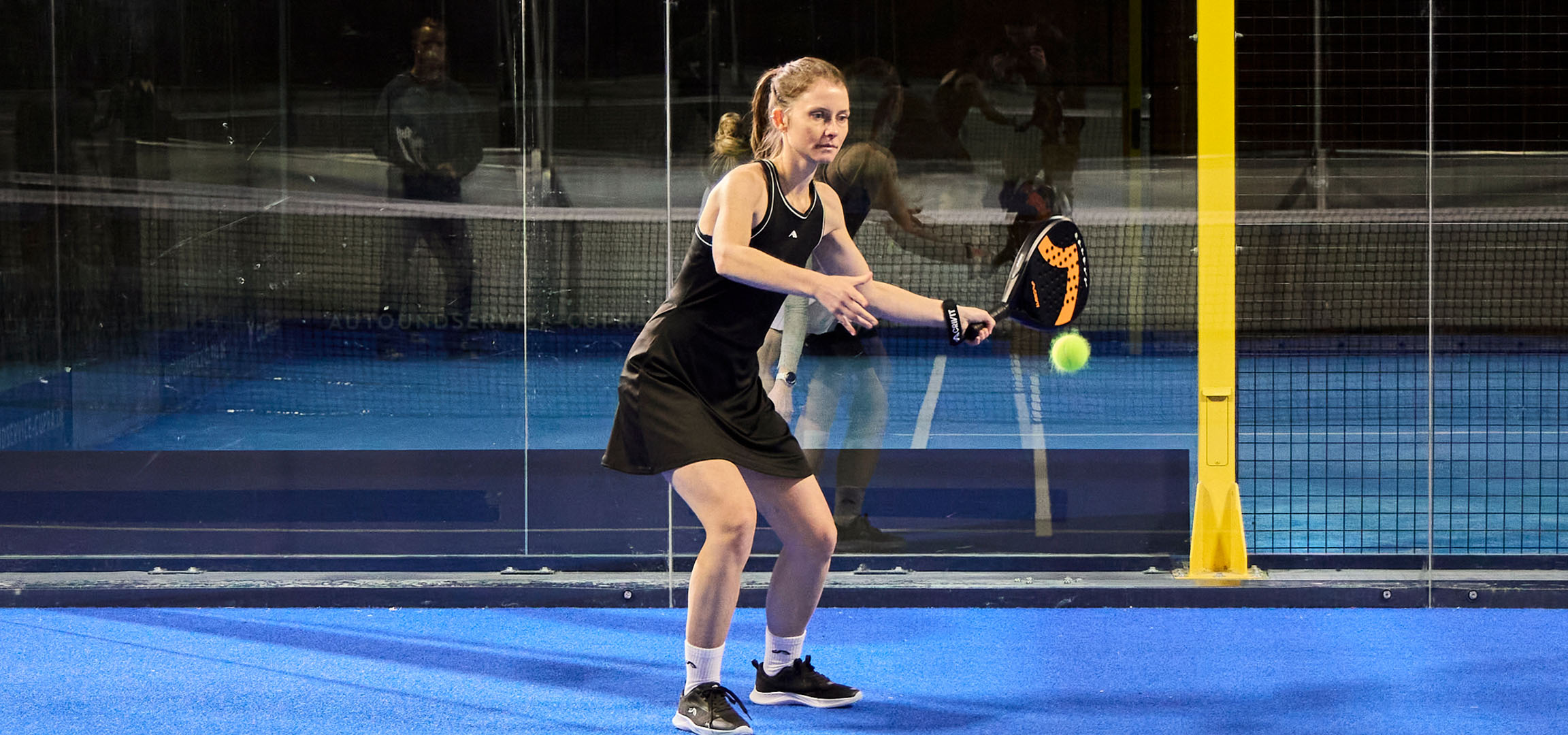 Femme jouant au padel sur un terrain bleu, vêtue de vêtements de sport noirs.