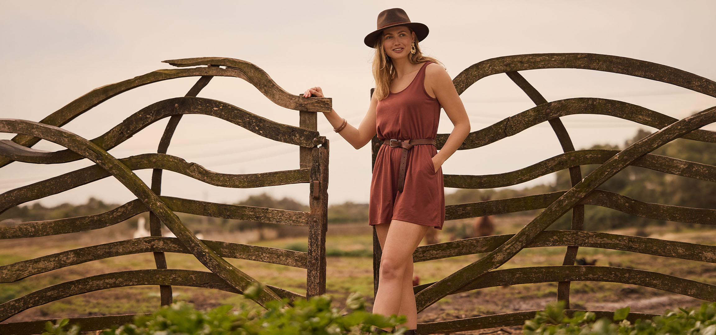 Femme en robe et chapeau, appuyée sur une clôture en bois à la campagne.