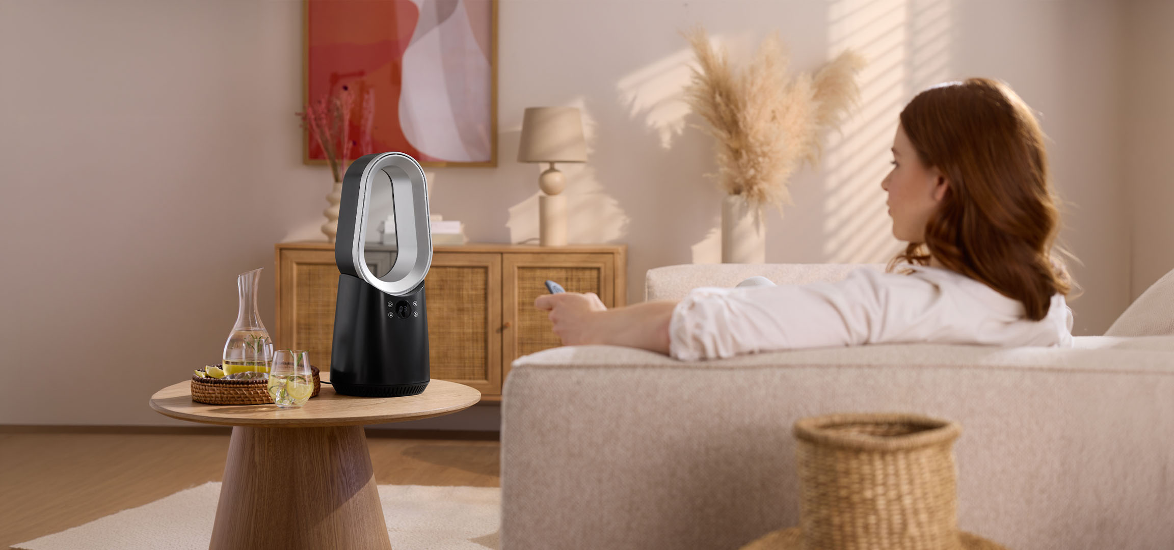 Femme sur canapé avec télécommande, ventilateur moderne et boissons sur table.