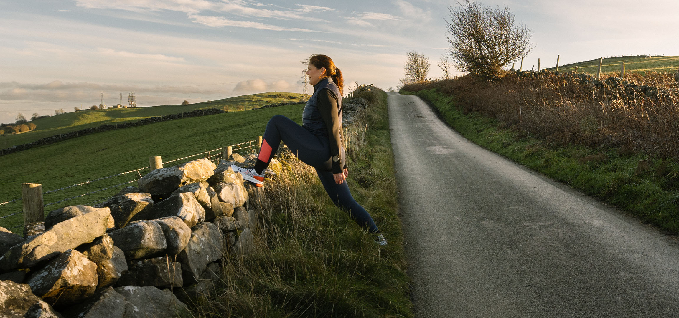 Femme s'étirant près d'un mur en pierre sur une route de campagne au coucher du soleil.