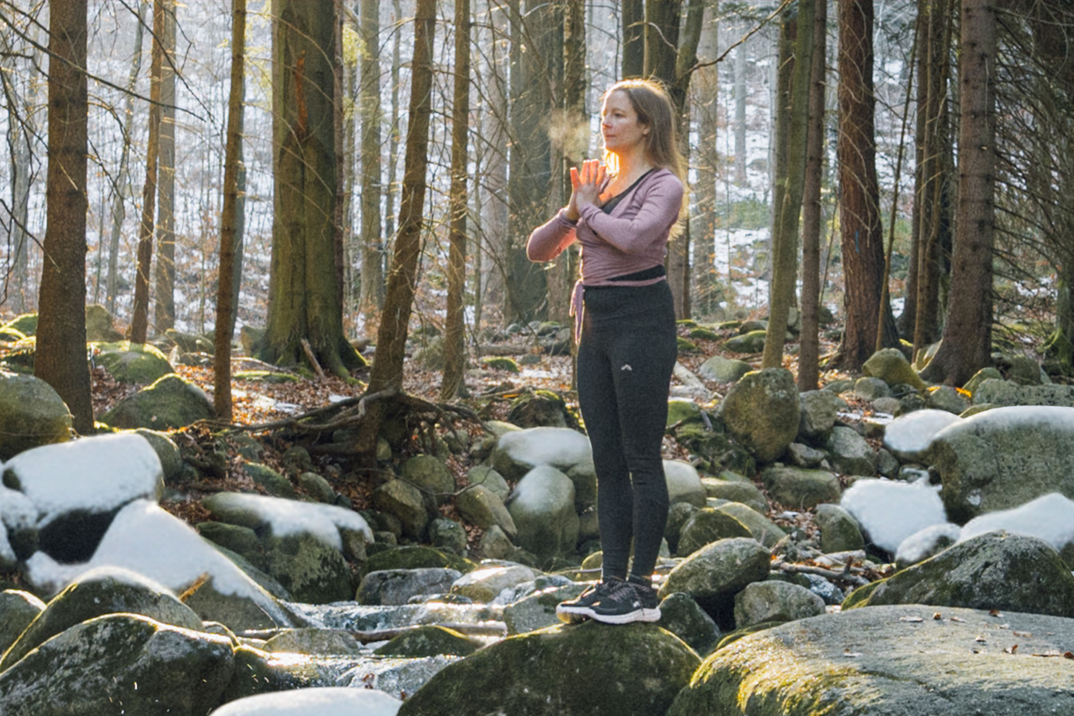 Femme en tenue de sport faisant du yoga dans une forêt avec des rochers et des arbres enneigés.