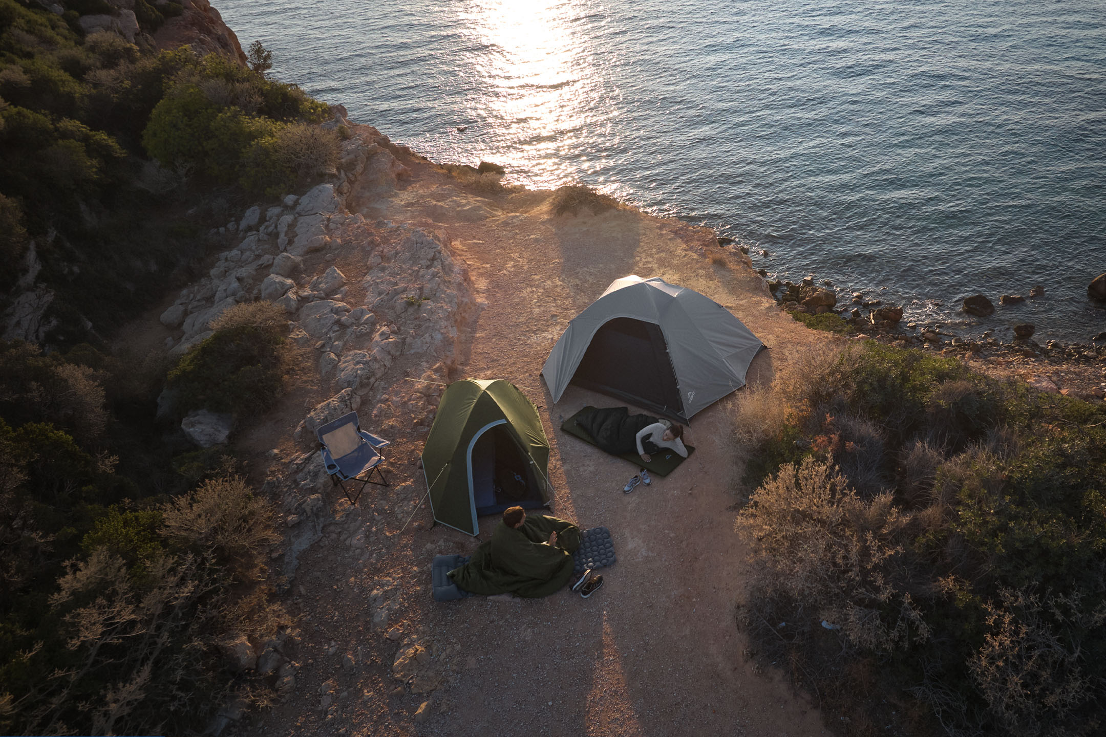 Deux tentes et sacs de couchage au bord de la mer au coucher du soleil, avec des campeurs qui se reposent.