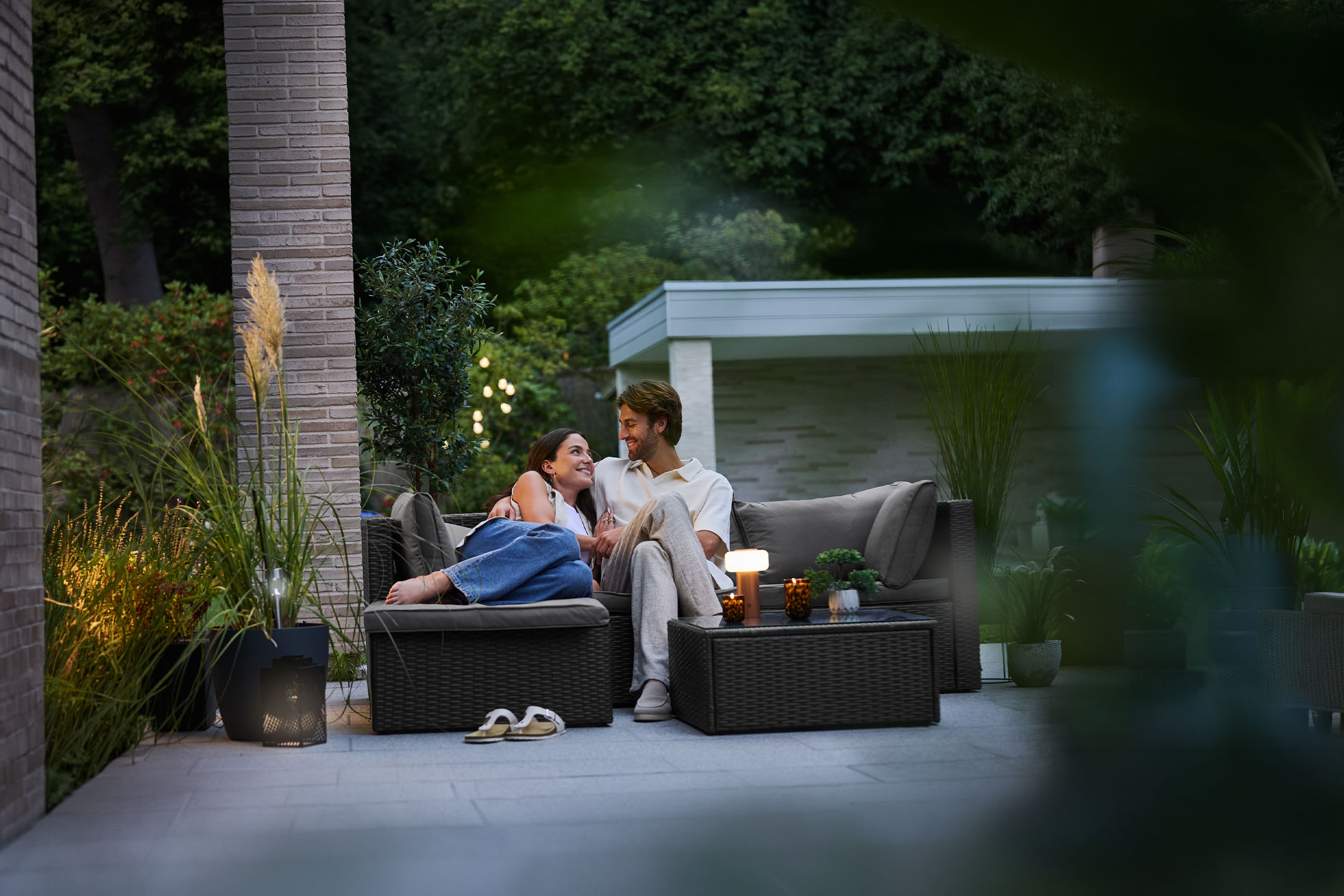 Couple se relaxant sur des meubles de jardin en rotin, éclairés par des guirlandes et des bougies.