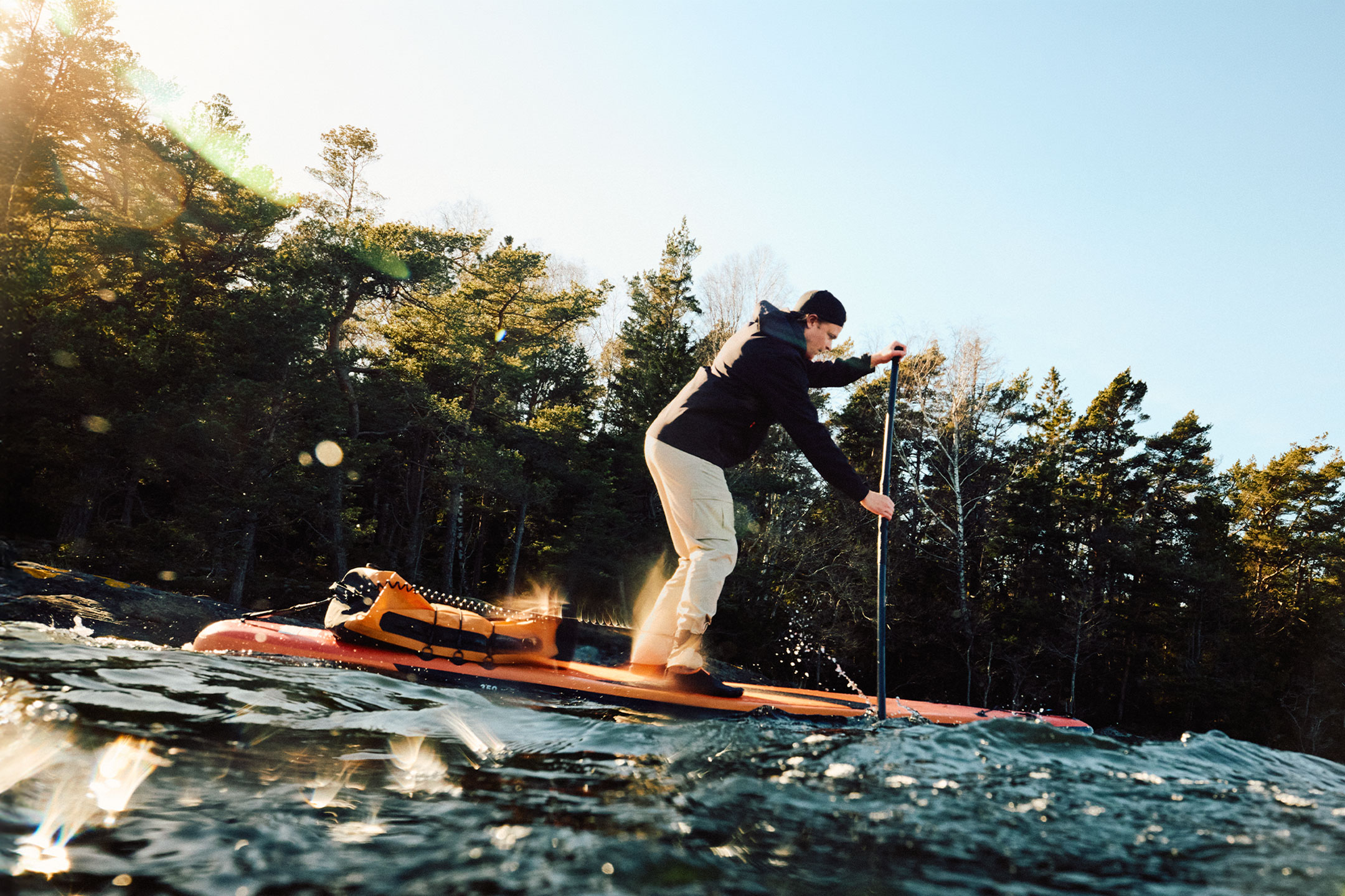 Homme en pantalon beige et veste noire faisant du paddle sur une planche orange
