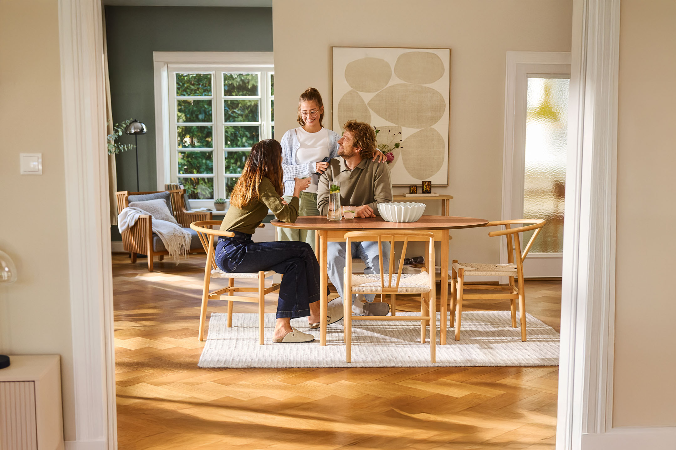 Trois personnes discutent autour d'une table en bois dans un salon lumineux avec de grandes fenêtres.