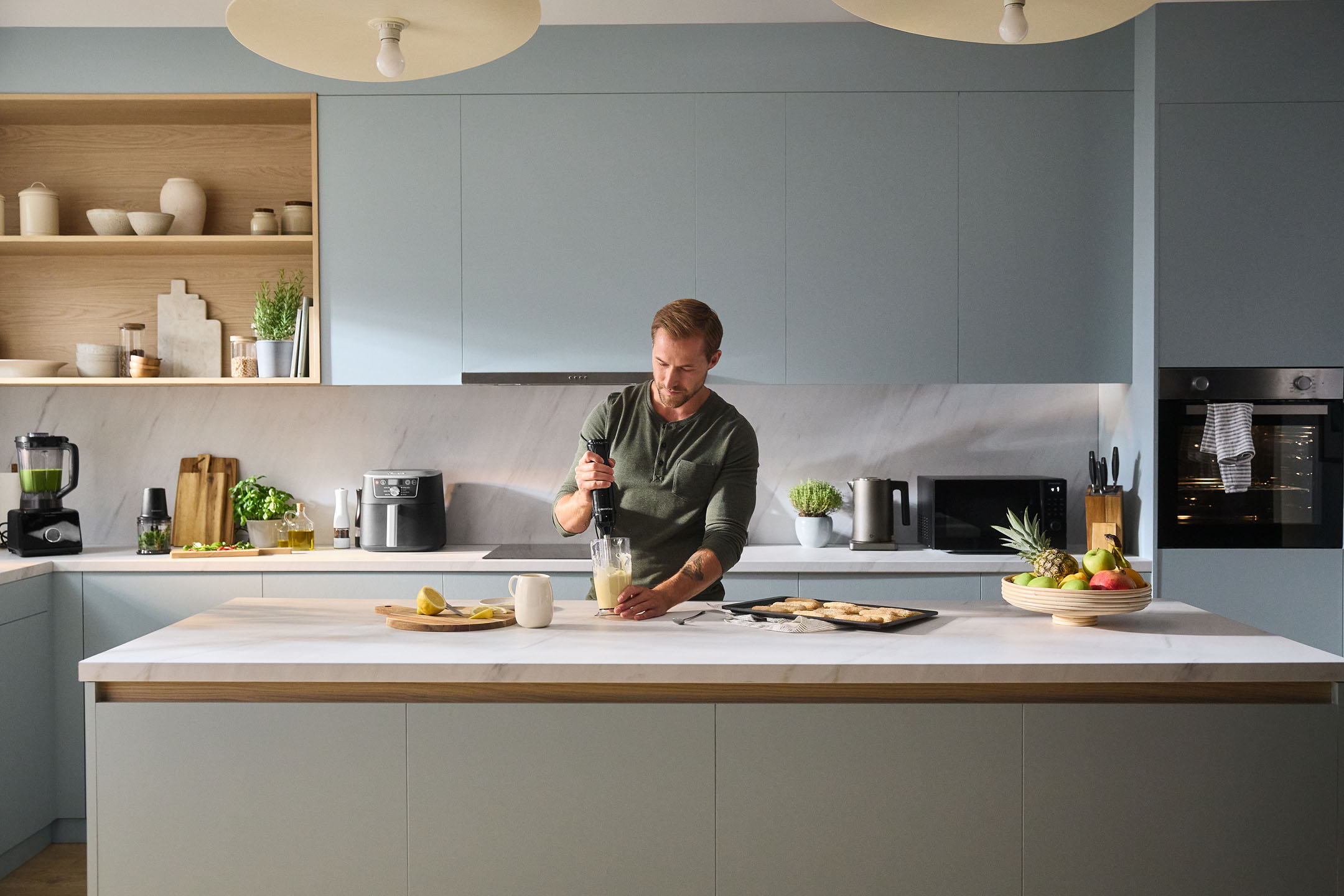 Homme utilisant un mixeur plongeant dans une cuisine moderne avec des appareils et des fruits.