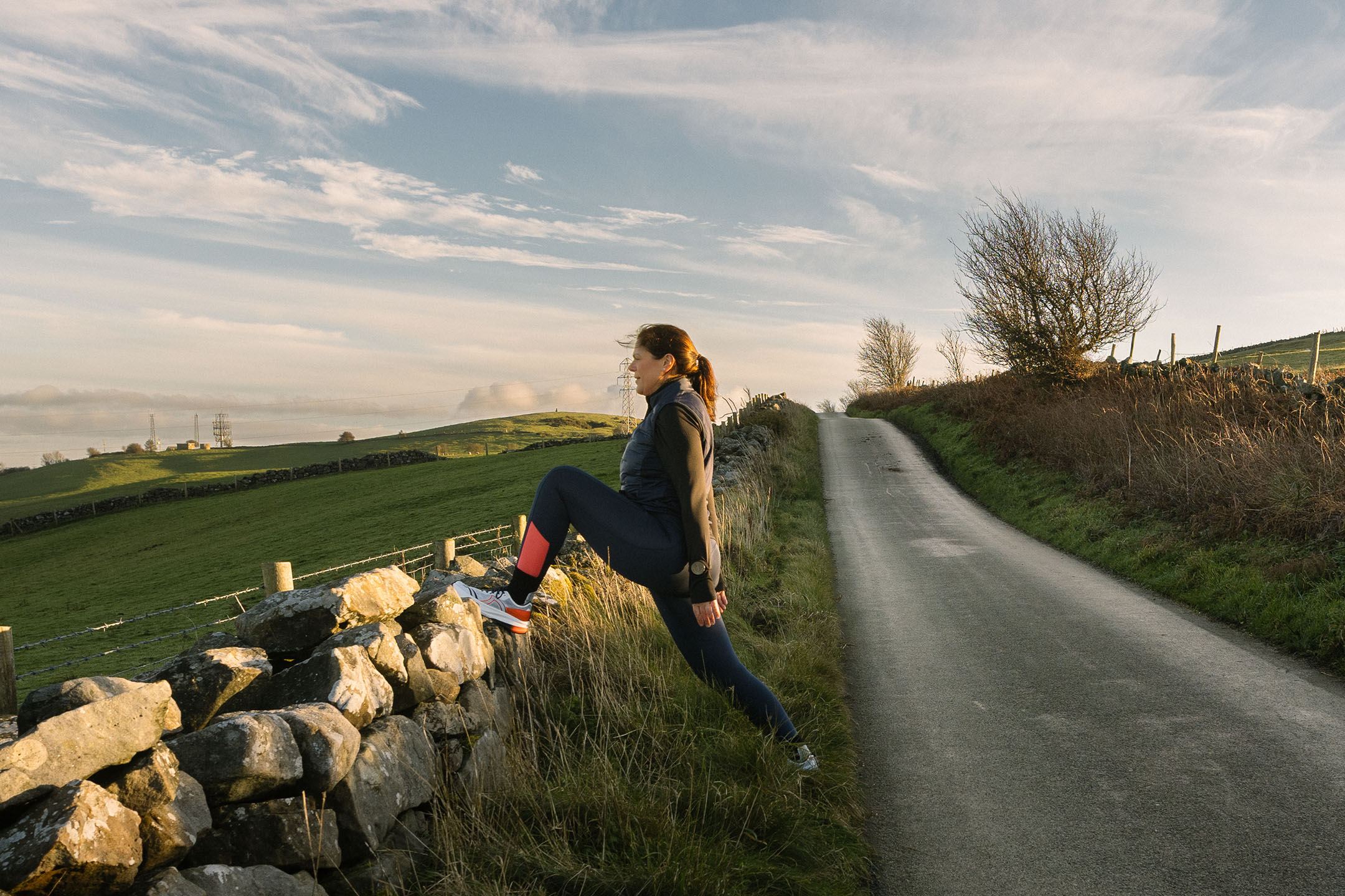 Femme en tenue de sport s'étirant sur un mur en pierre au bord d'une route de campagne.