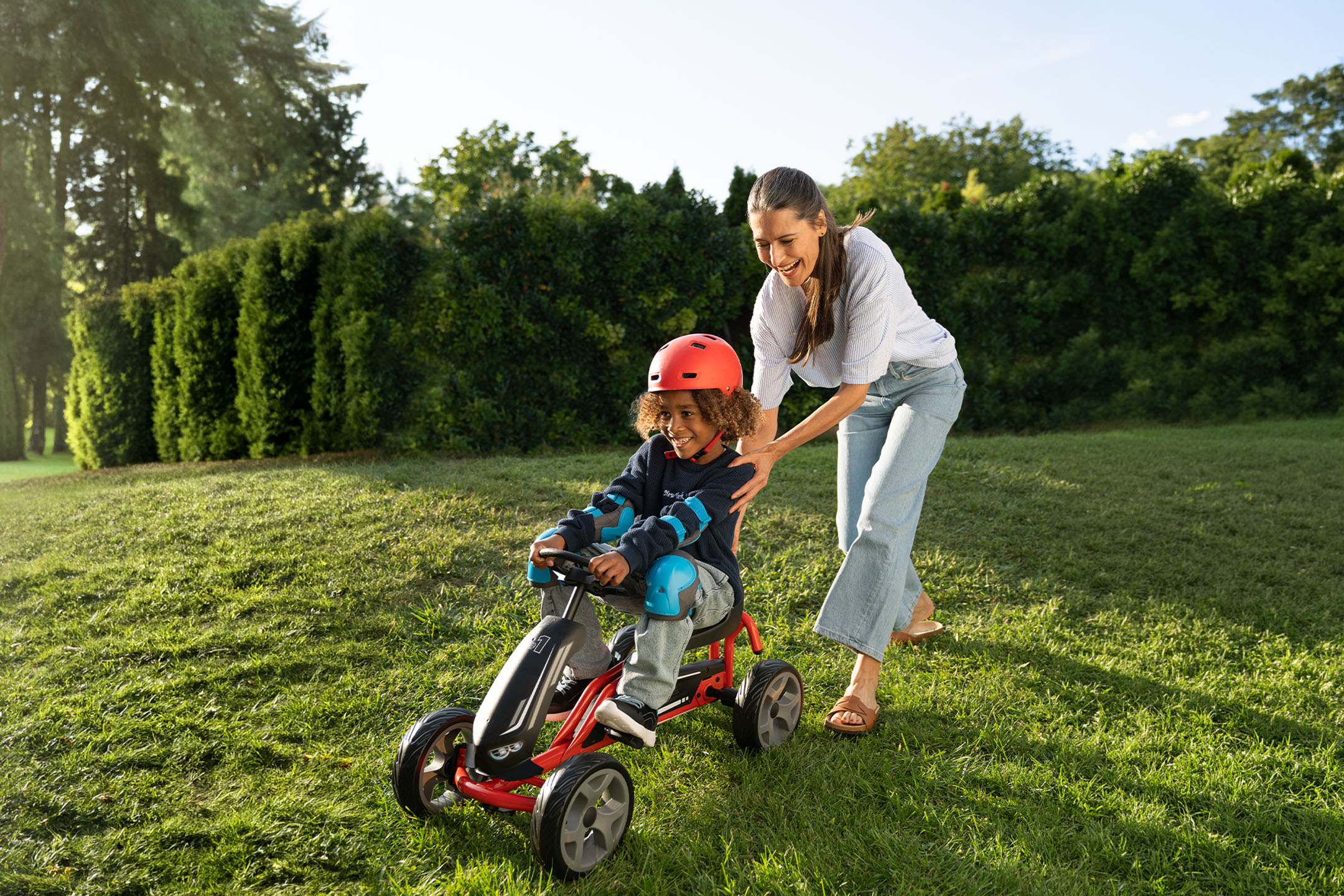 Mère et enfant jouant dehors avec un kart à pédales, l'enfant portant casque et protections.