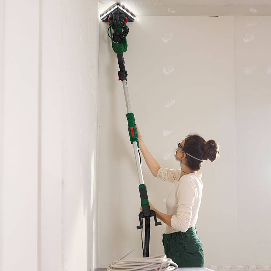 Femme ponçant un mur avec une ponceuse murale télescopique dans un coin.