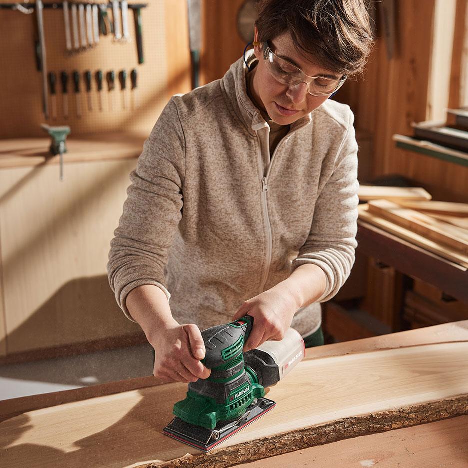 Femme avec ponceuse électrique ponçant un morceau de bois dans un atelier.