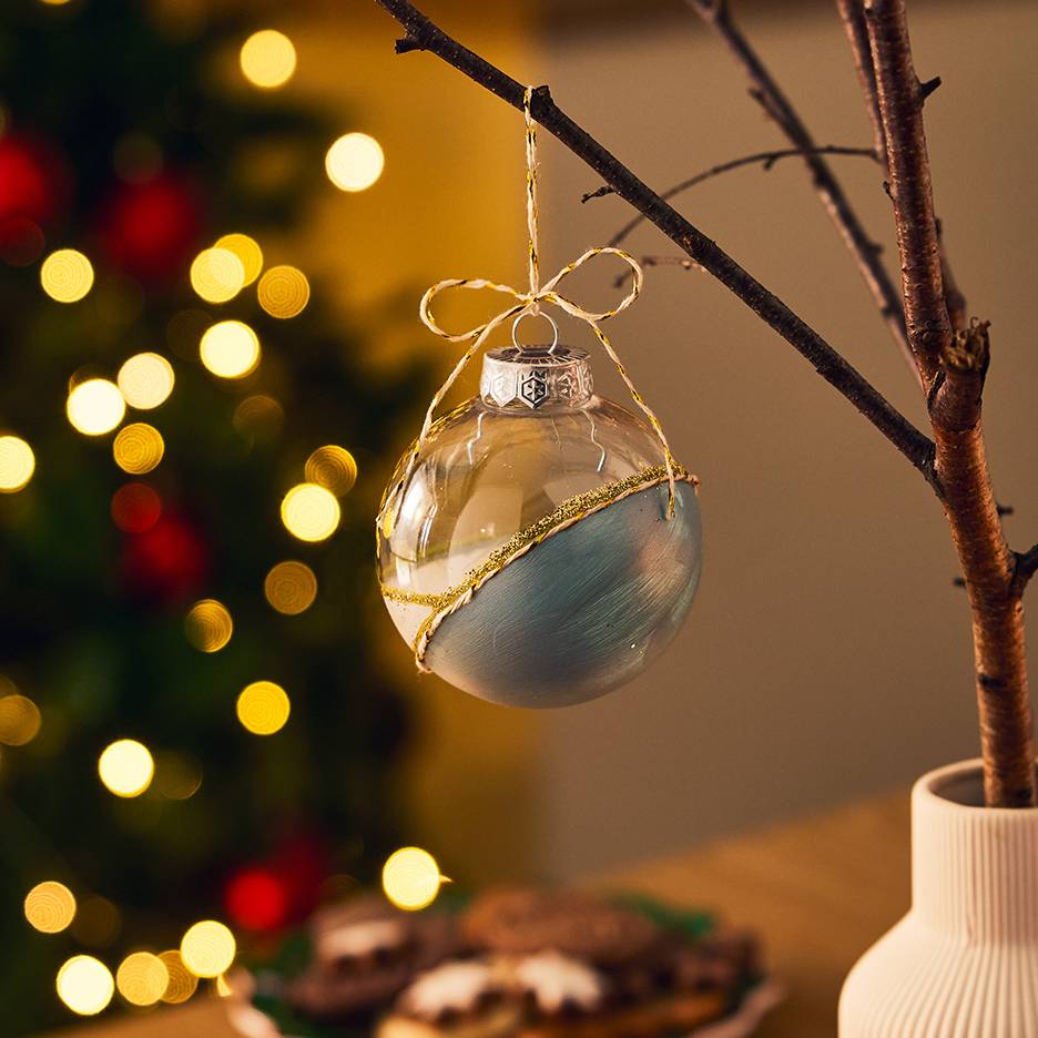 Boule de Noël transparente et bleue avec des paillettes dorées, suspendue à une branche avec des lumières de Noël en arrière-plan.