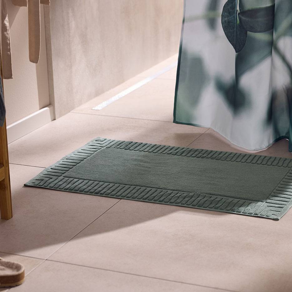 Tapis de bain vert texturé sur carrelage clair, à côté d'un rideau de douche.