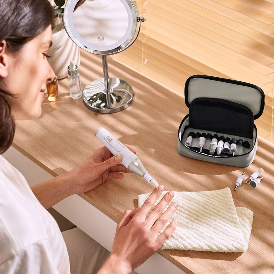 Femme utilisant une lime à ongles électrique avec accessoires, à côté d'un miroir et d'une trousse.