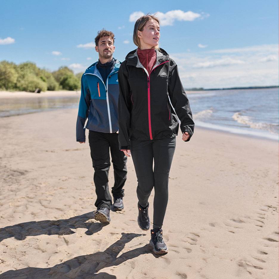 Couple en vestes et pantalons de randonnée marchant sur une plage de sable au bord de la mer.