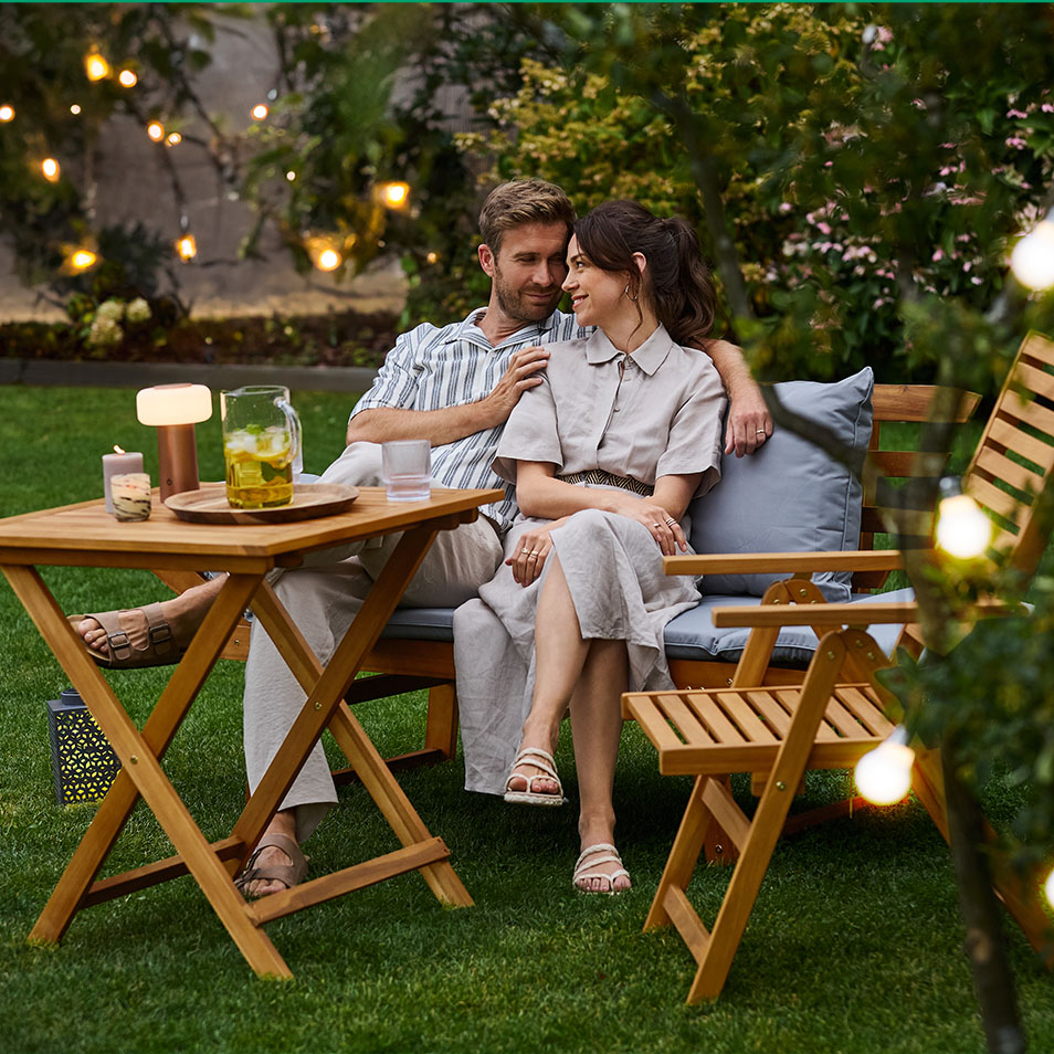 Couple se relaxant sur un banc de jardin en bois avec coussins, table et chaises assorties.