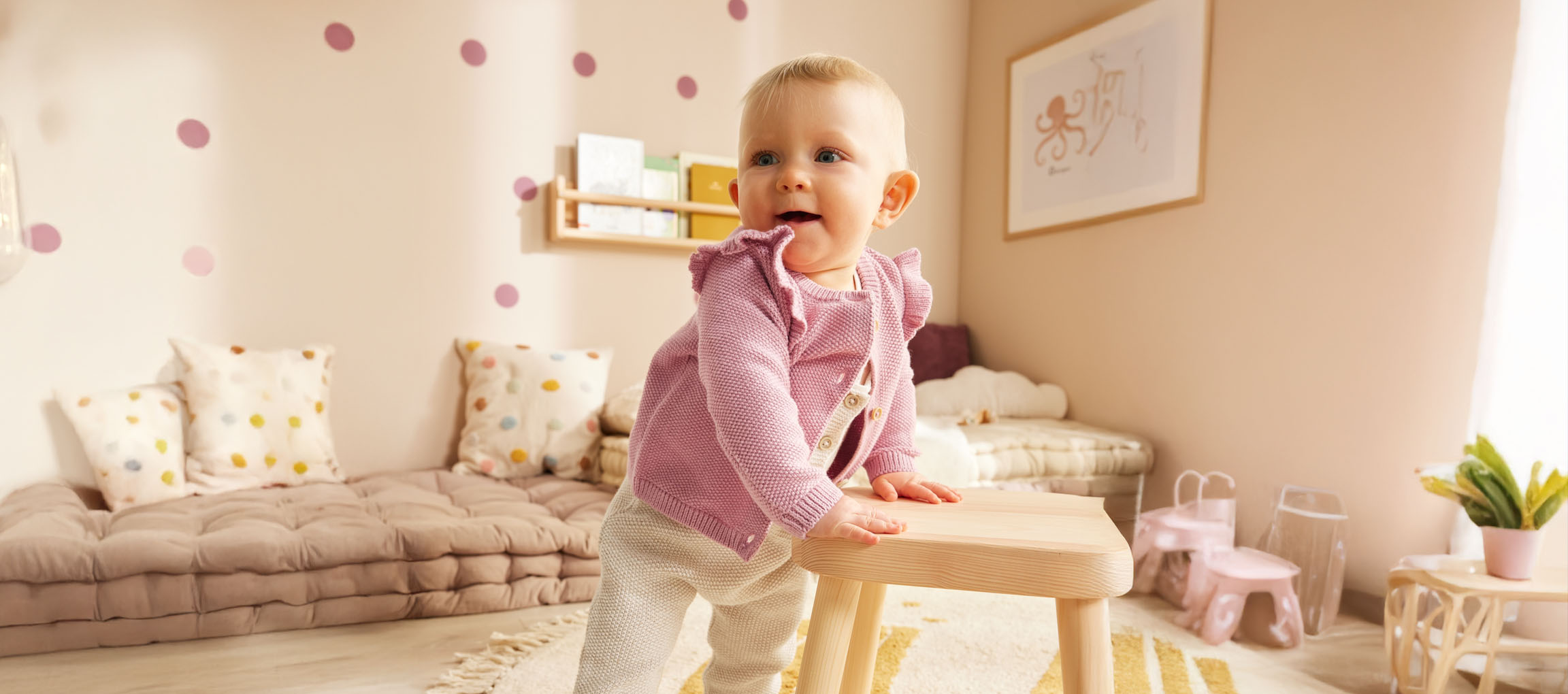 Un bébé dans un joli cardigan rose se tient debout près d'un tabouret en bois dans sa chambre d'enfant.