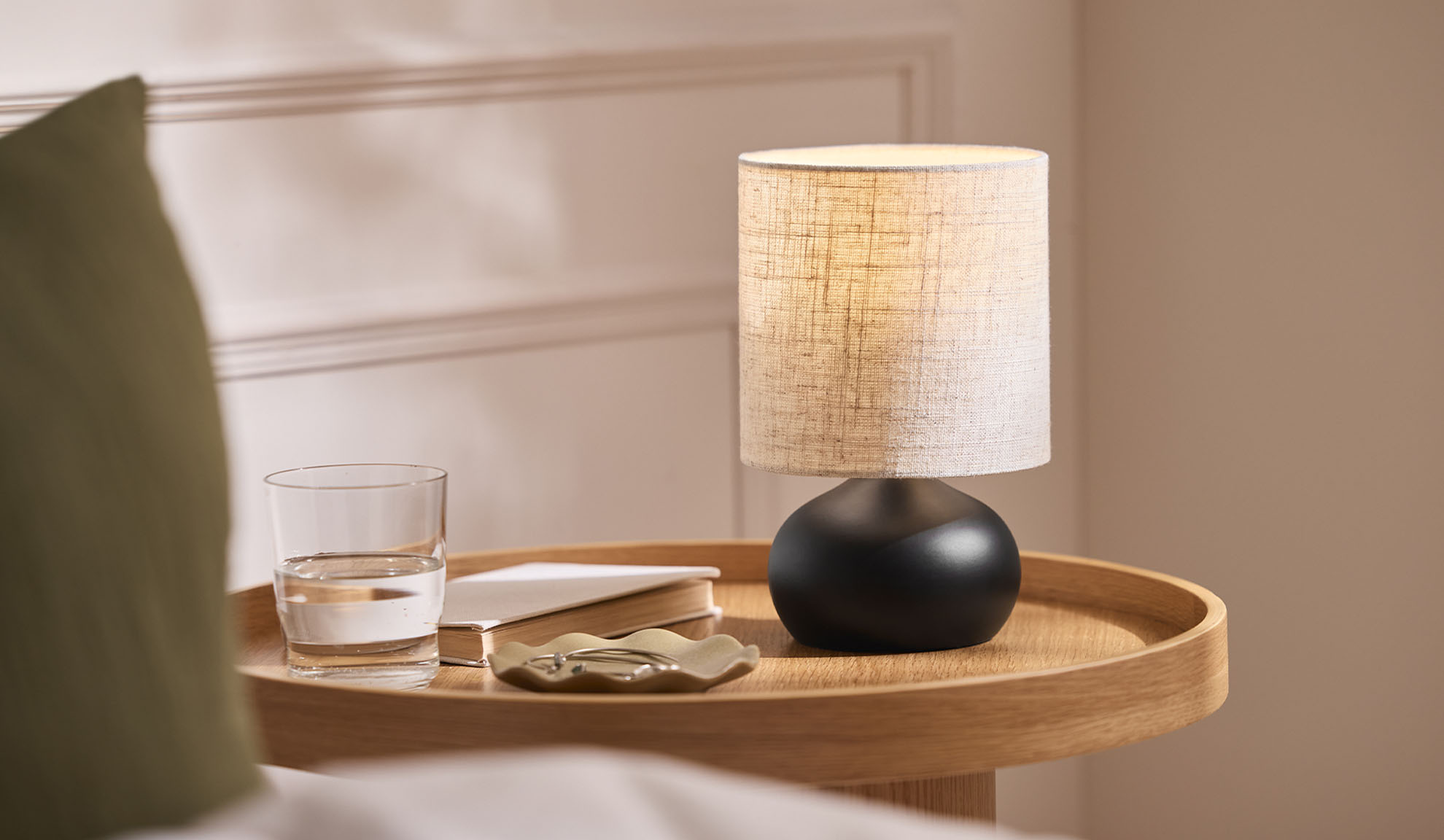Lampe de chevet, verre, livre et plateau à bijoux sur un plateau en bois à côté du lit.