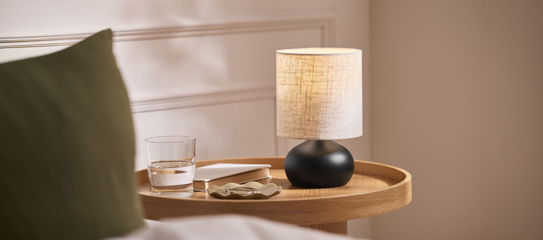 Lampe de chevet avec abat-jour en tissu, verre d'eau et livre sur un plateau en bois.
