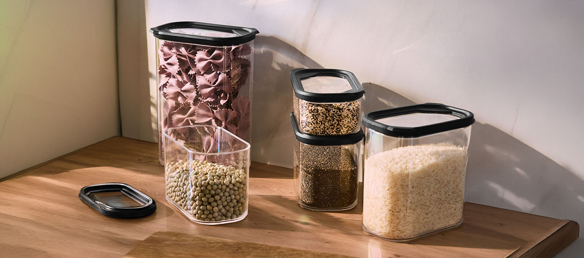 Récipients de cuisine transparents avec couvercles noirs, remplis de pâtes, lentilles, quinoa, graines de chia et riz sur une surface en bois.