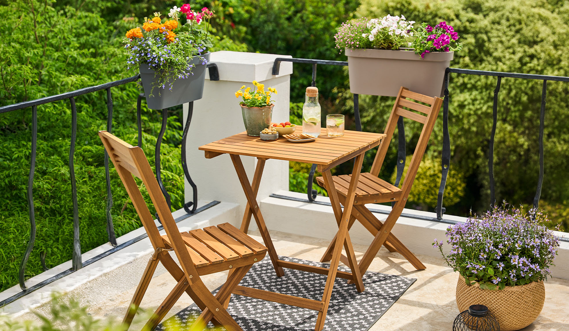 Balcon avec ensemble bistrot en bois, fleurs en pot et boissons sur un tapis à motifs.