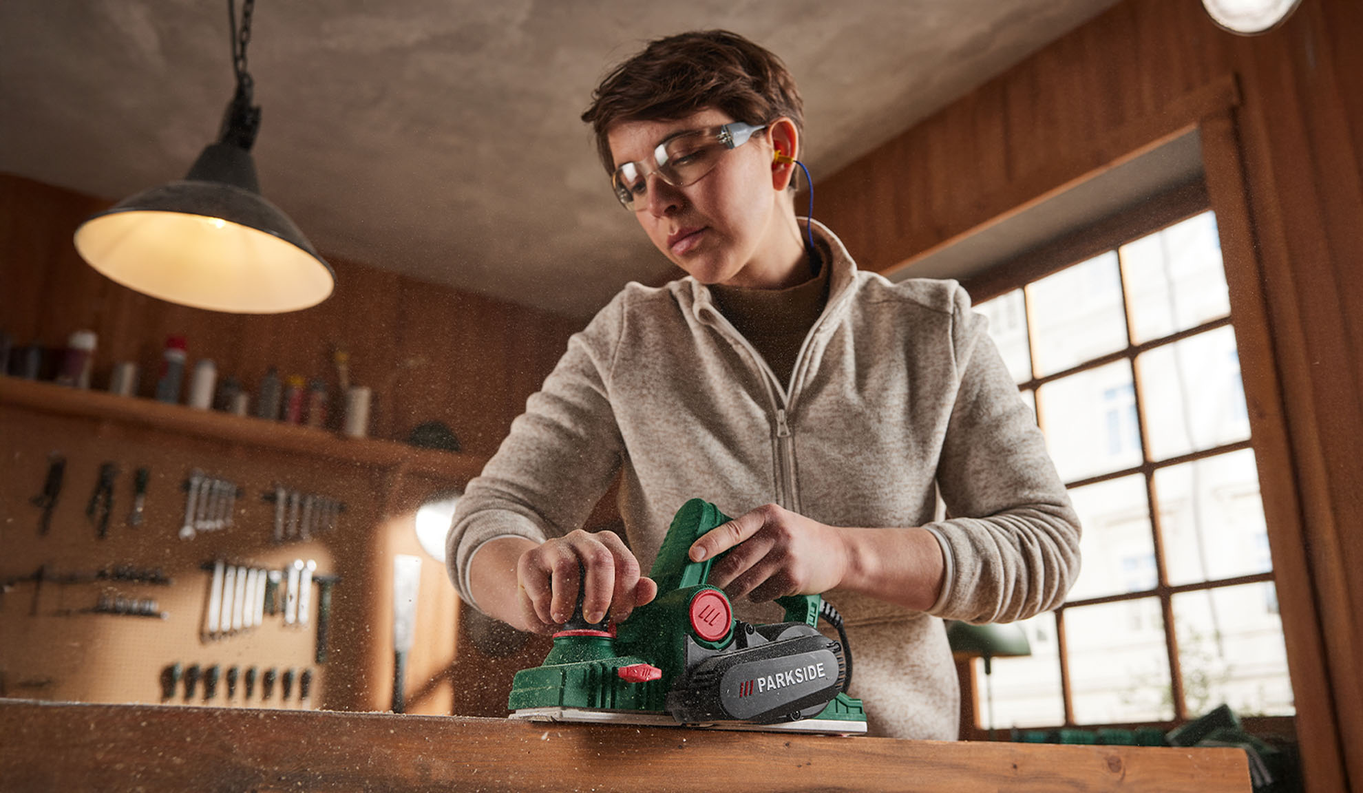 Femme avec lunettes de protection et bouchons d'oreille utilisant une raboteuse électrique Parkside sur du bois.
