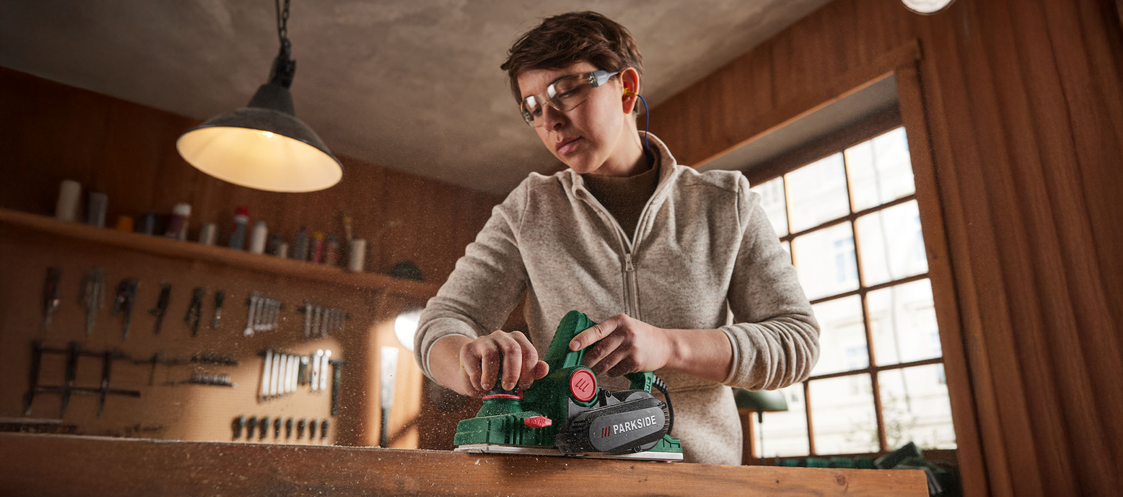 Personne utilisant une raboteuse électrique Parkside sur du bois, avec des lunettes de sécurité et des bouchons d'oreille.