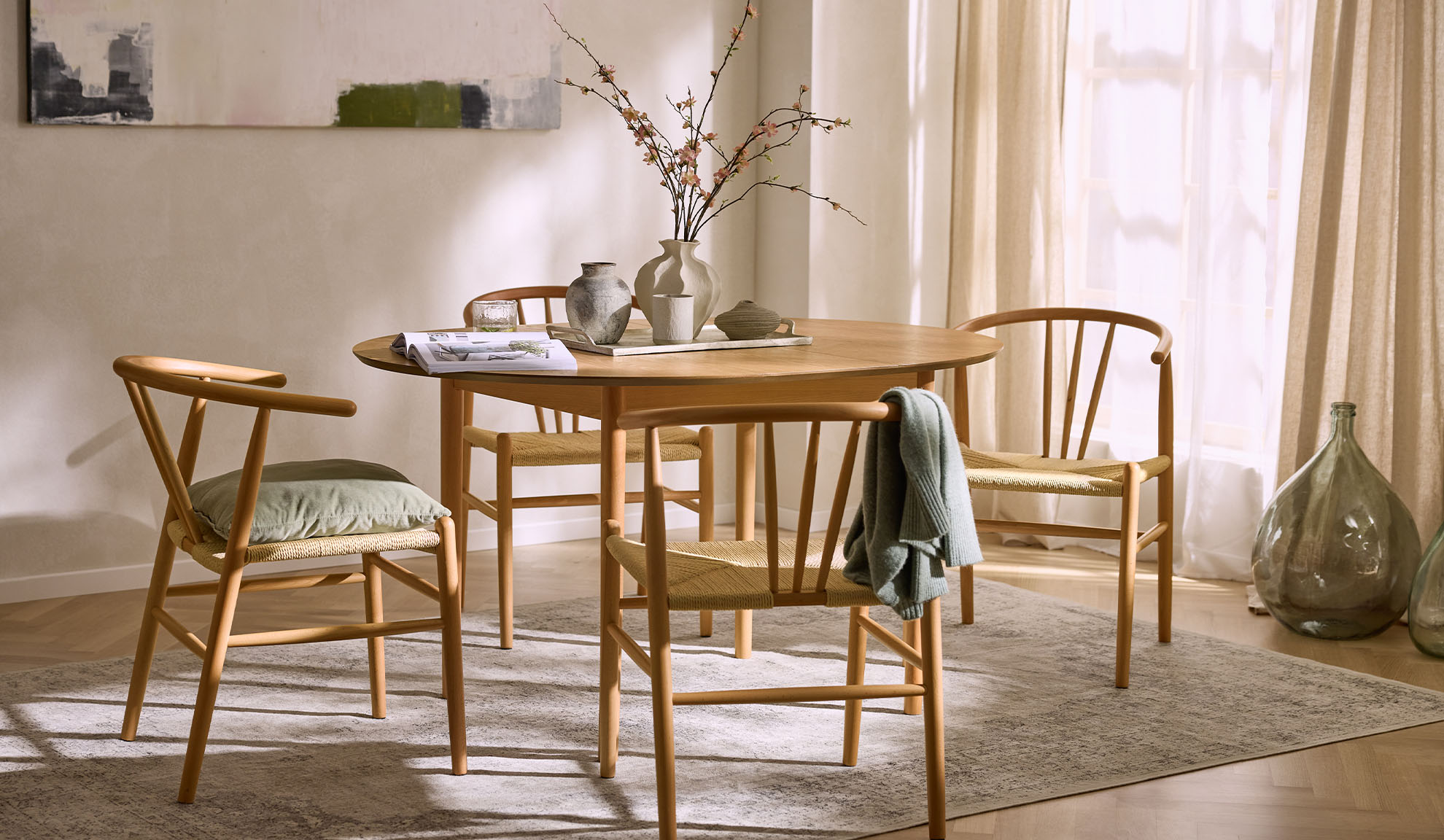 Salle à manger lumineuse avec table ronde en bois, chaises en bois avec assises en corde et décor floral.