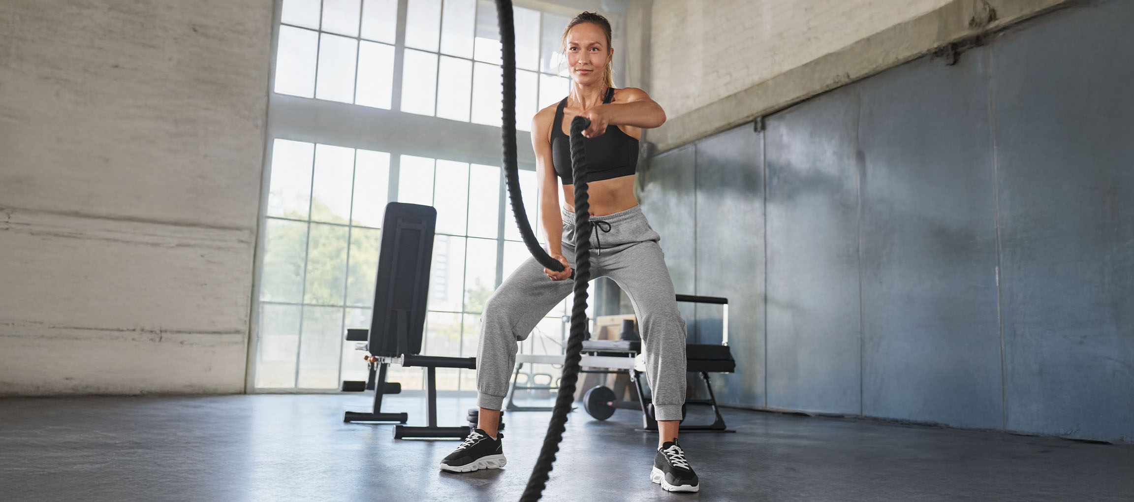 Femme en brassière de sport noire et pantalon de survêtement gris s'entraînant avec des cordes de combat
