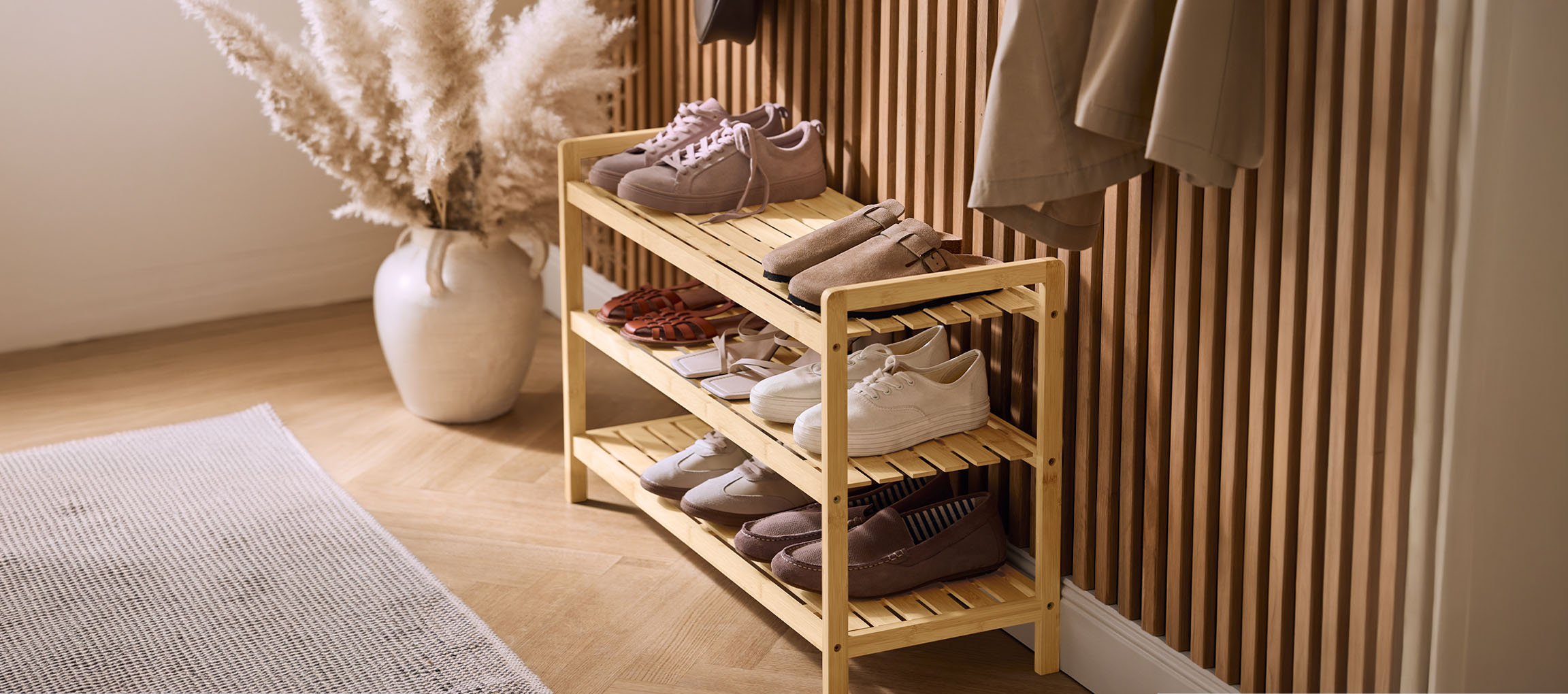 Étagère à chaussures en bois avec diverses chaussures, un vase d'herbe de la Pampa et un manteau au mur.