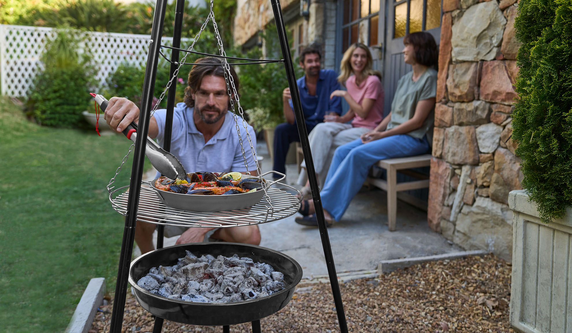 Homme grillant des fruits de mer sur un trépied, amis se relaxant en arrière-plan.