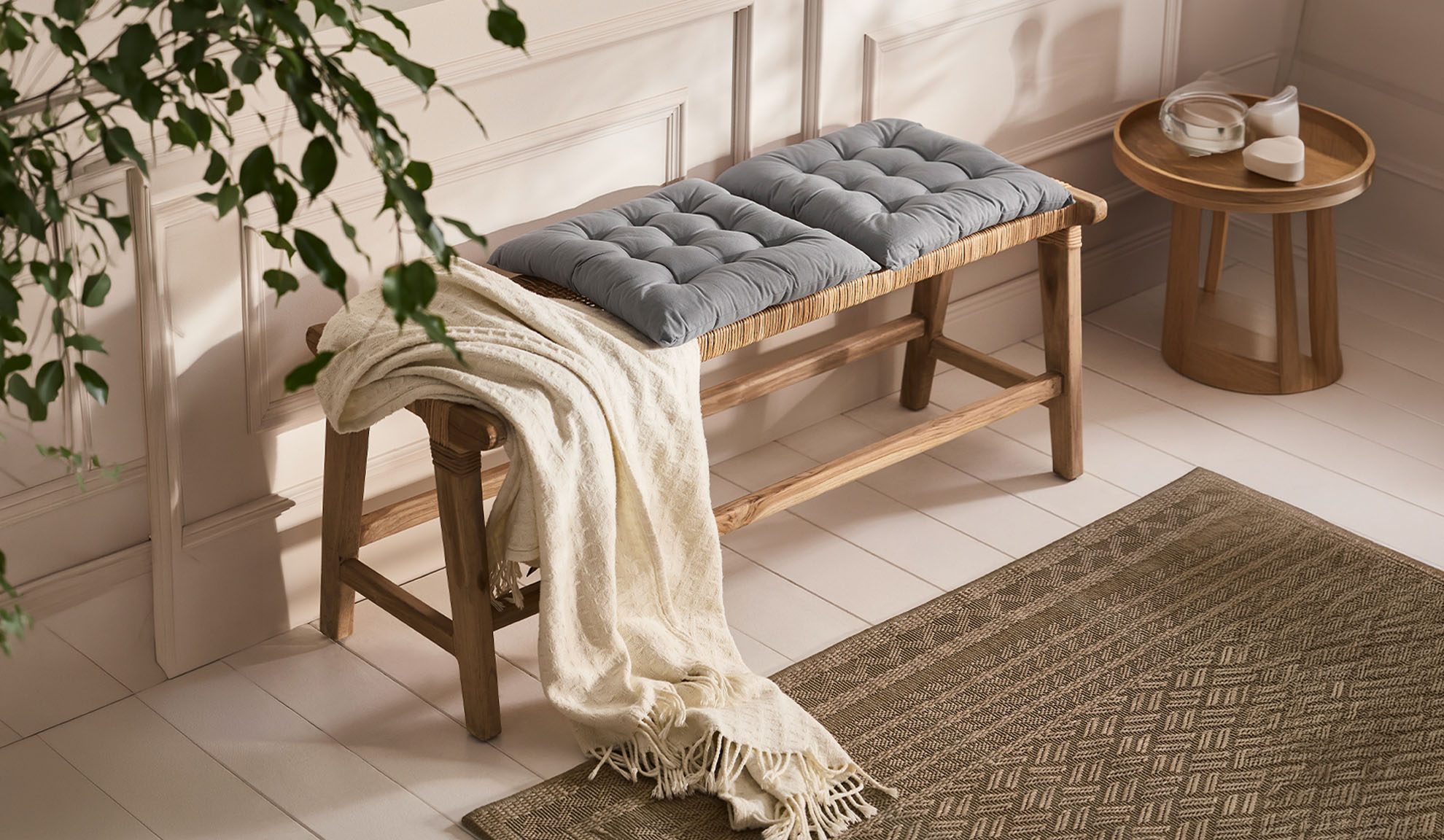 Banc en bois avec coussins gris et plaid, à côté d'une table en bois avec vaisselle, un tapis au sol.