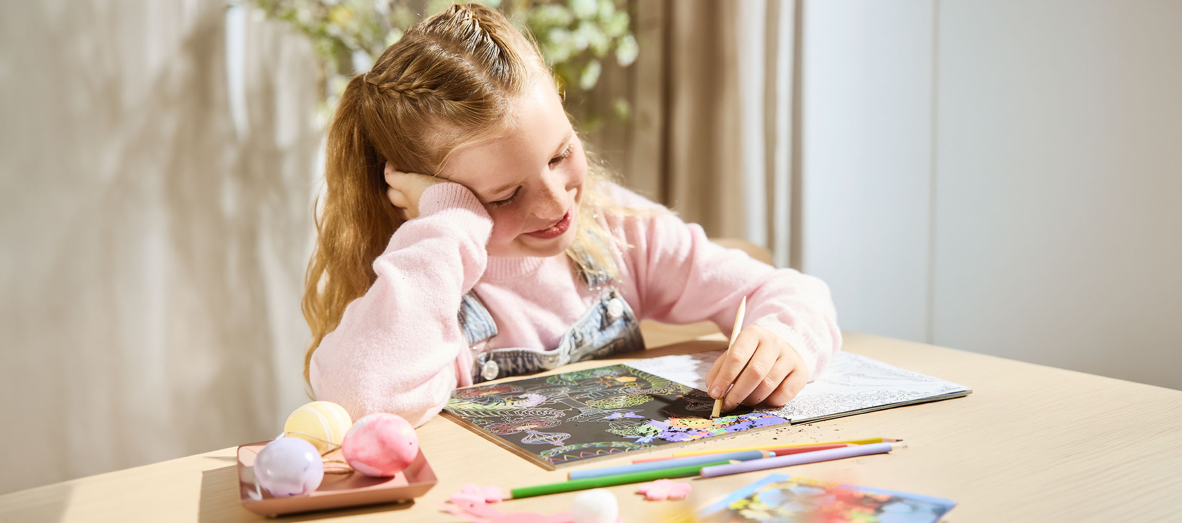 Une fille gratte un motif coloré sur du papier à gratter noir, avec des crayons de couleur à proximité.