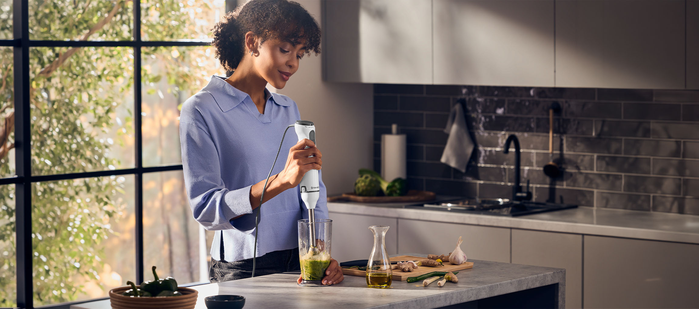 Femme dans la cuisine utilisant un mixeur plongeant pour faire un smoothie vert.