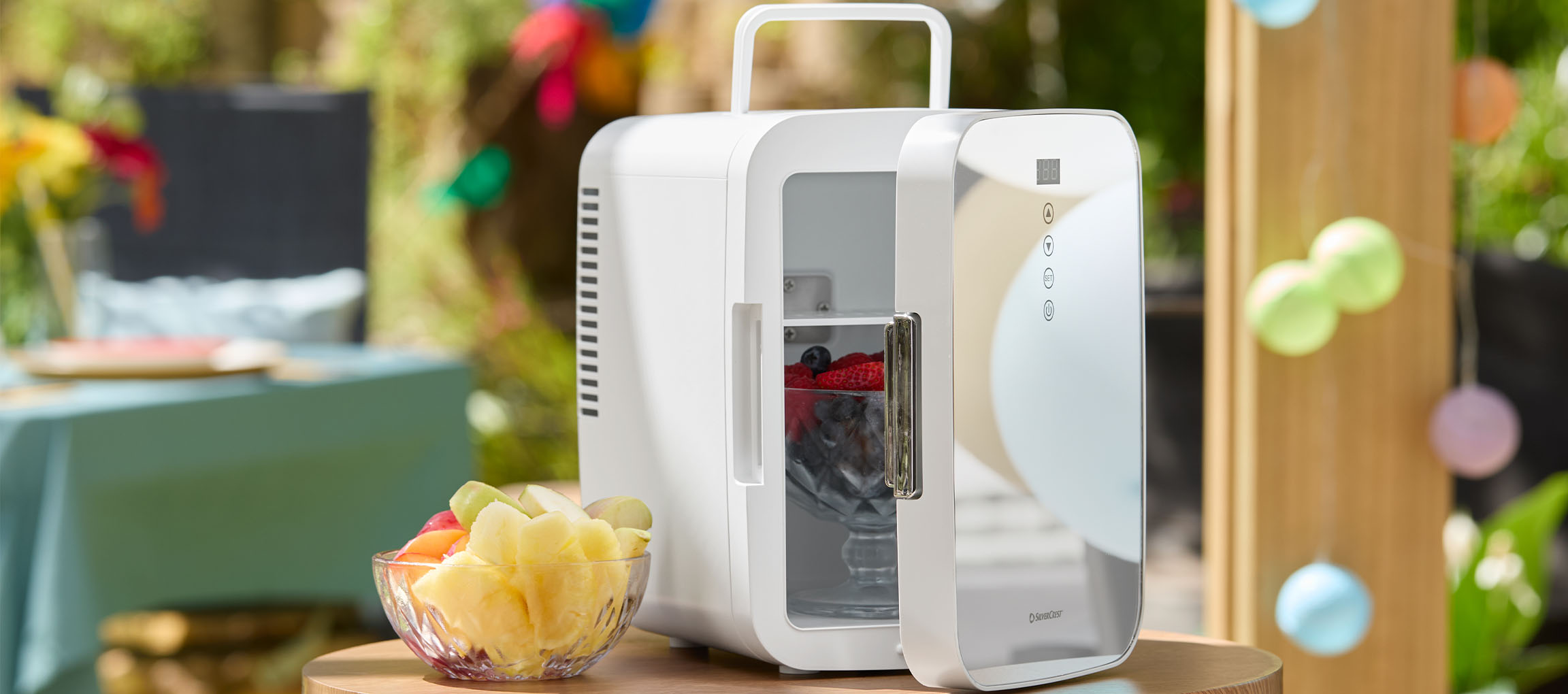 Mini-réfrigérateur blanc avec porte ouverte rempli de baies et un bol de fruits sur une table extérieure.