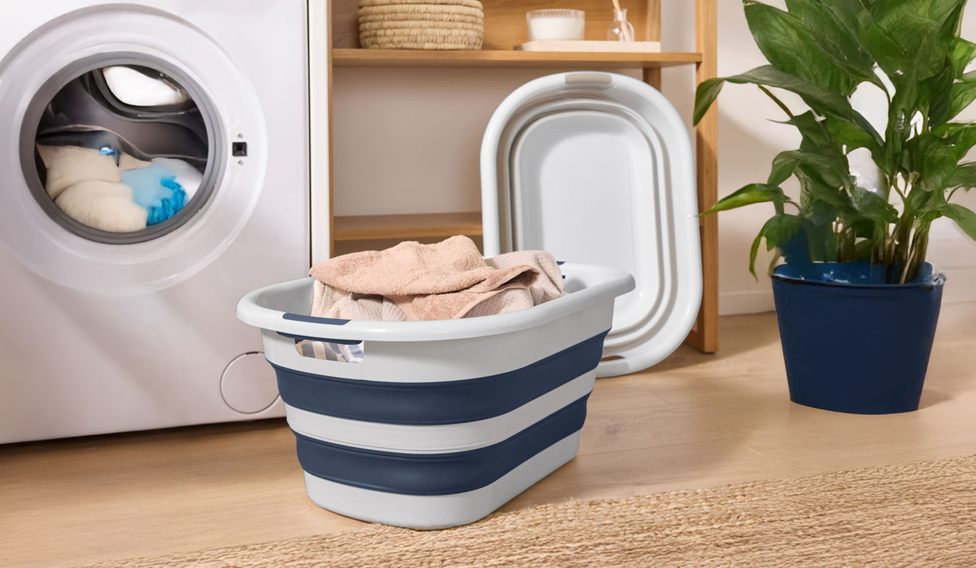 Une machine à laver avec des vêtements, un panier à linge pliable et une plante.