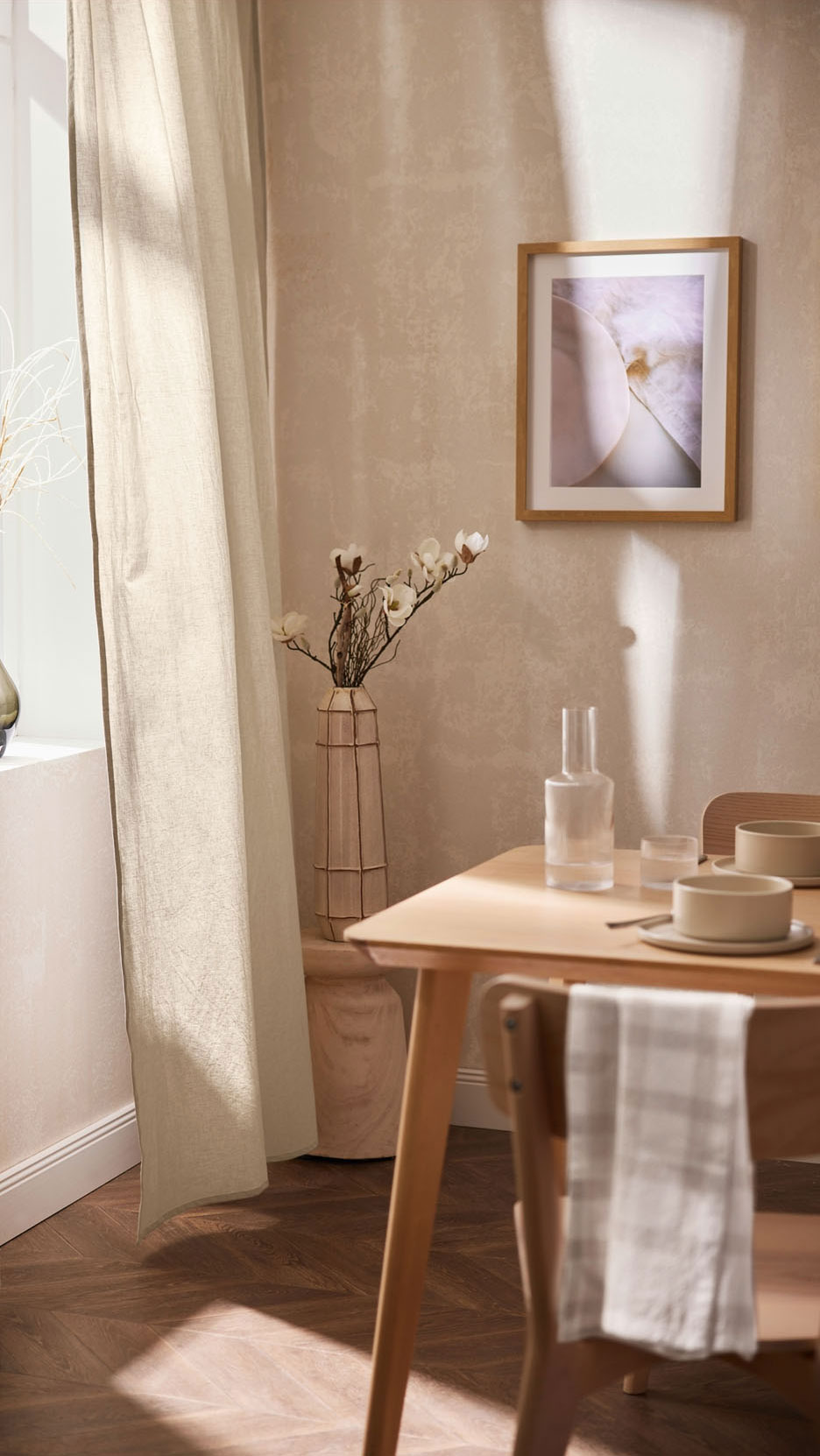 Une table en bois clair avec de la vaisselle et un vase de fleurs, près d'un rideau en lin beige.