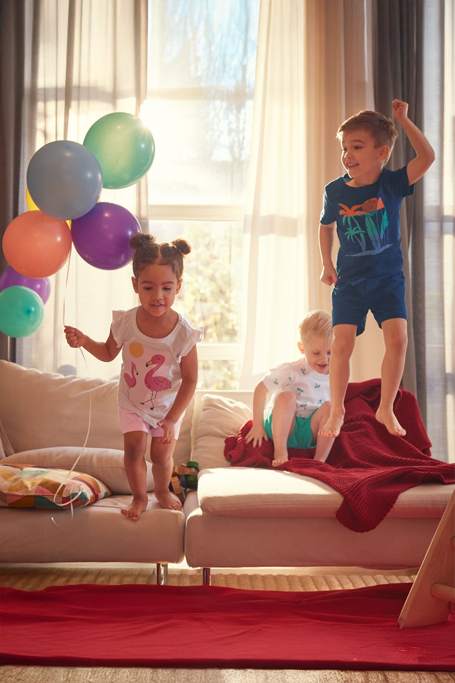 Trois enfants sautent sur un canapé, près d'un bouquet de ballons.
