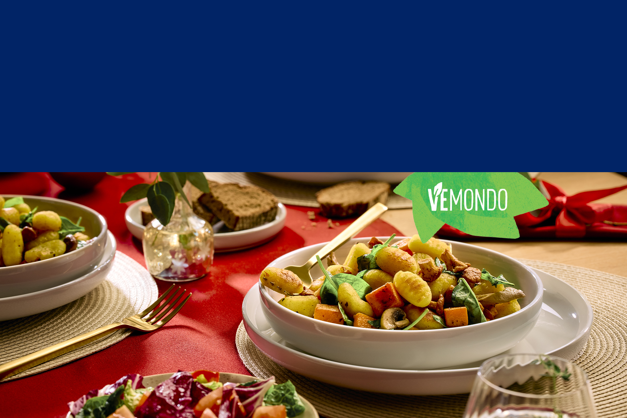 Plat de gnocchis aux légumes, salade et pain sur une table rouge, avec le logo Vemondo.