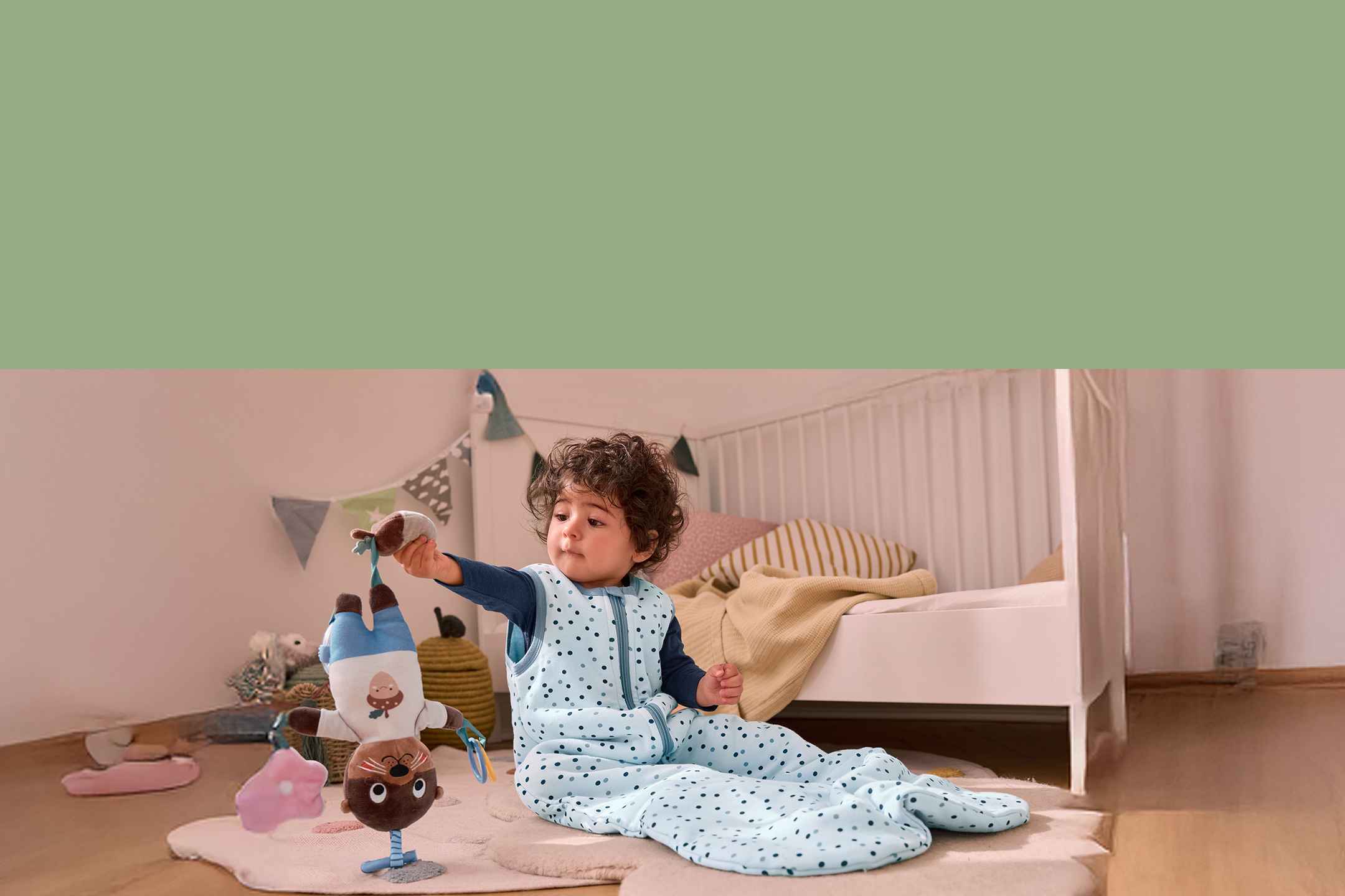 Un bébé en gigoteuse à pois bleus joue avec une peluche dans une chambre d'enfant.