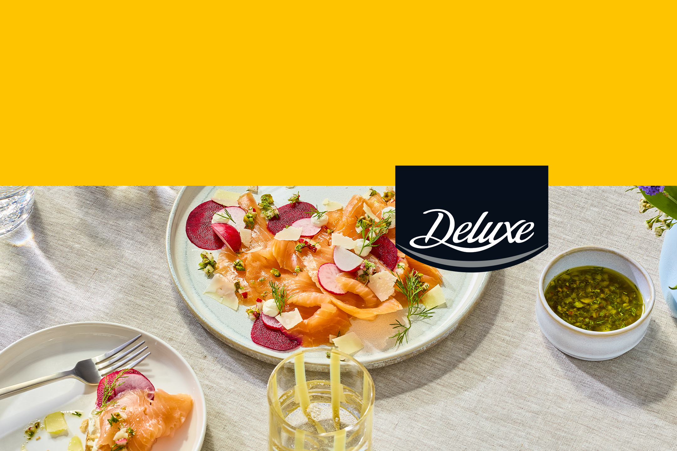 Carpaccio de saumon fumé avec radis, betterave et sauce verte, sur une table avec le logo 'Deluxe'.