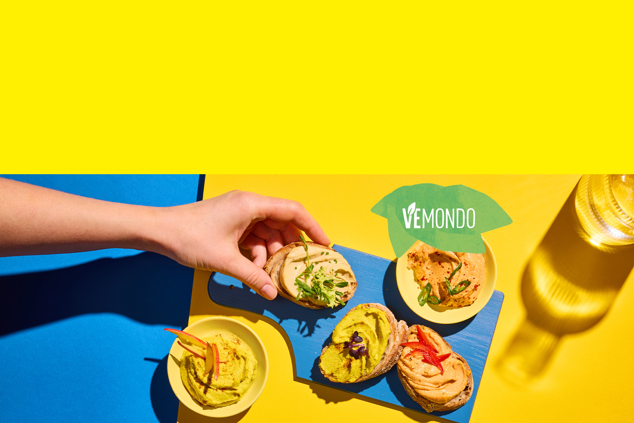 Une main prend une tartine de houmous sur une planche bleue, avec d'autres tartines et bols de houmous.