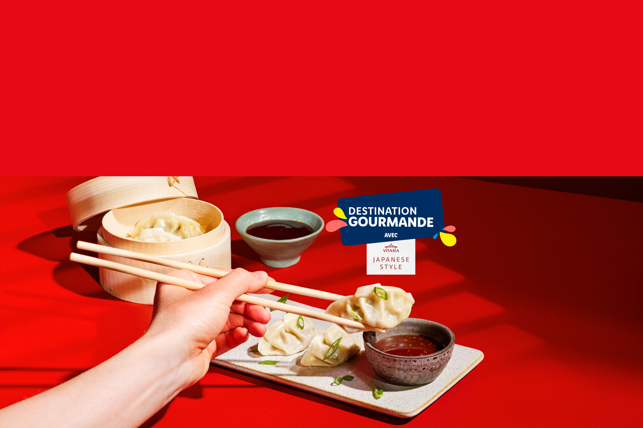 Une main tenant des baguettes avec des raviolis japonais, un cuit-vapeur en bambou et des sauces sur une table rouge. Thème : Destination Gourmande, style japonais.