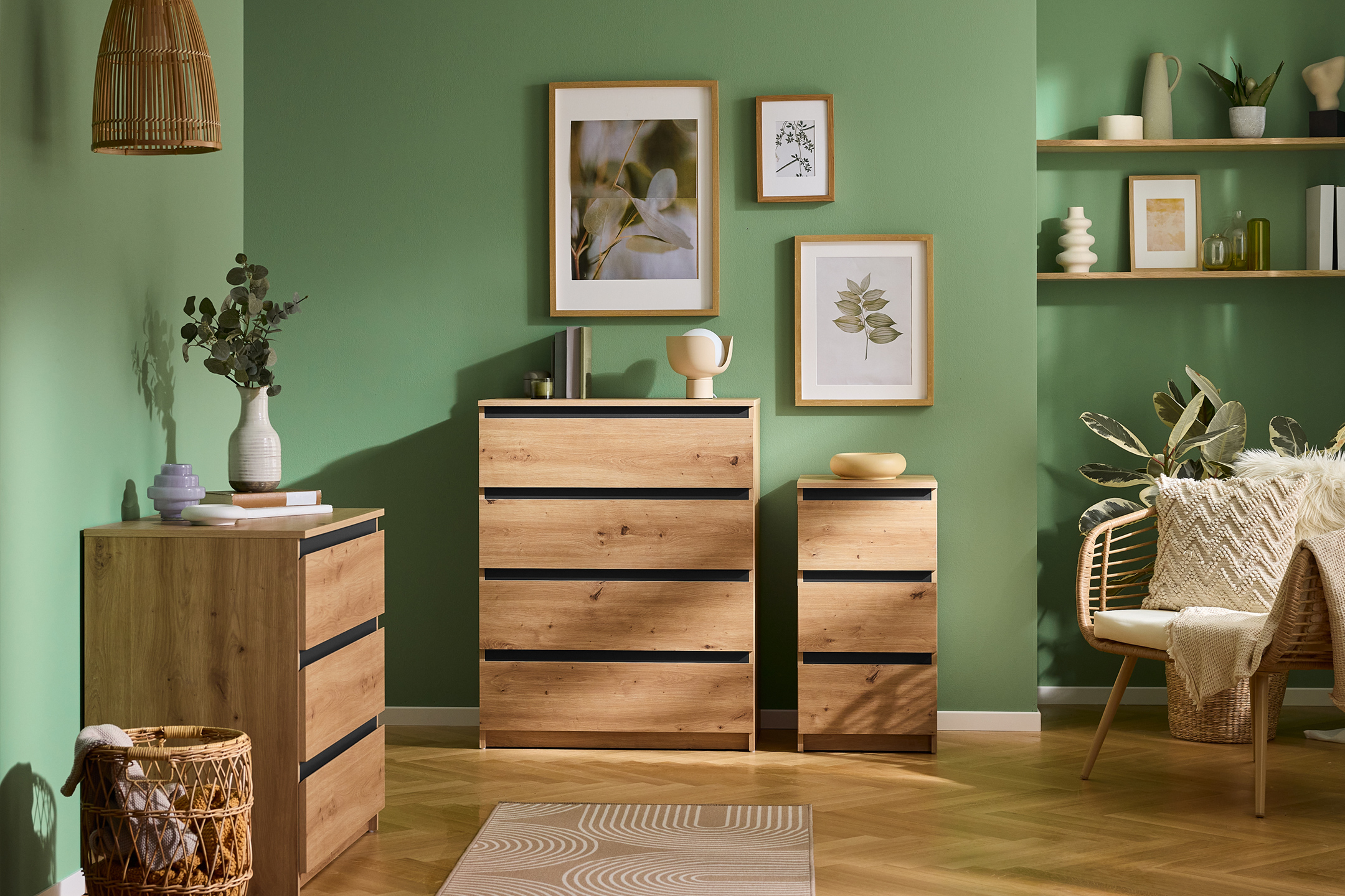 Commode en bois clair avec tiroirs noirs, étagères murales et fauteuil en rotin dans une pièce verte.